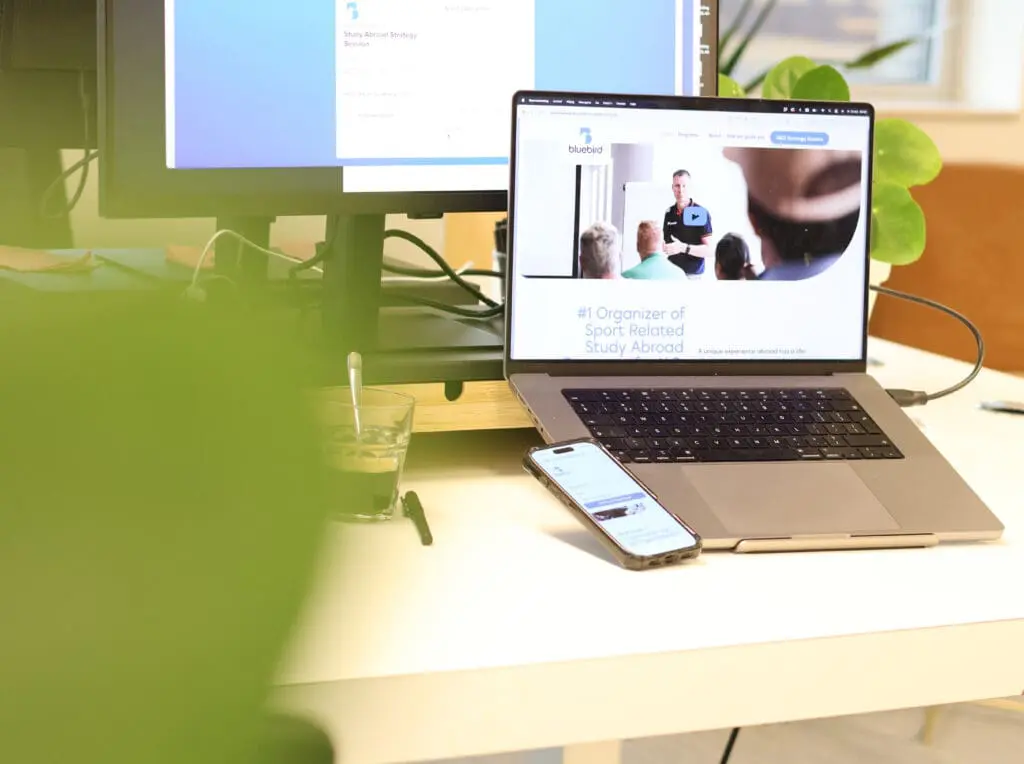 Werkplek met laptop waarop de Bluebird-website zichtbaar is, naast een smartphone en monitor op een bureau.