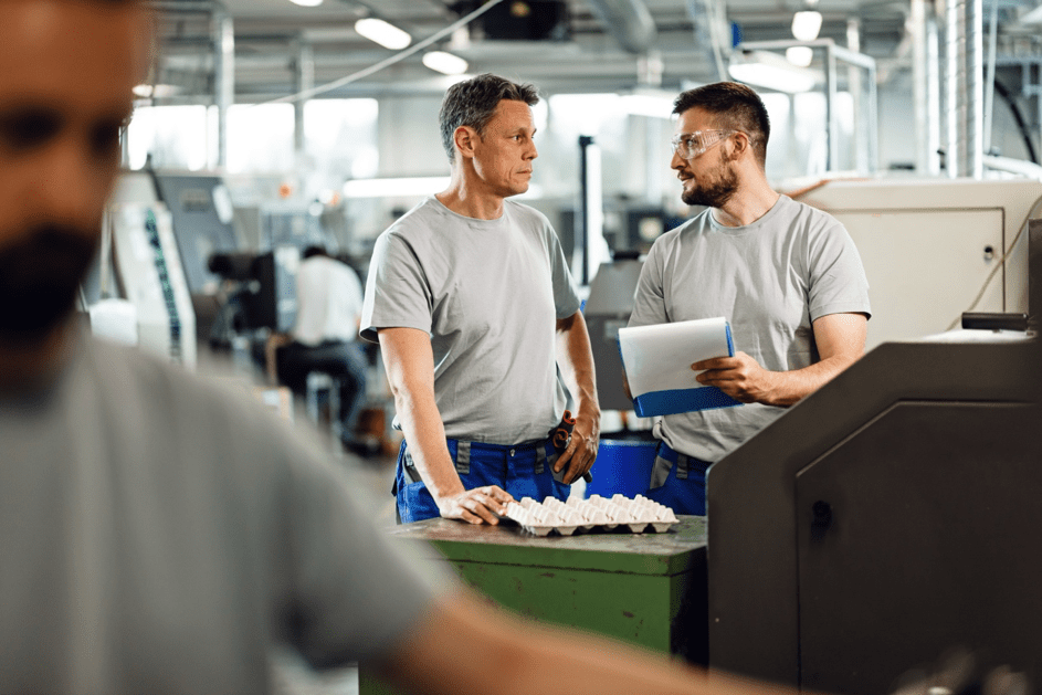 Werknemers in een fabriek praten bij een machine terwijl één van hen een clipboard met notities vasthoudt.