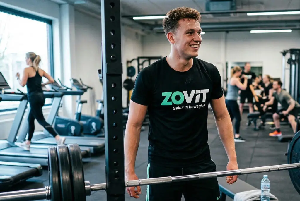 Man in sportschool met Zovit shirt bij gewichtsstang, met sporters en loopbanden op de achtergrond.