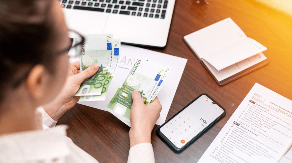 Persoon telt eurobankbiljetten aan bureau met laptop en smartphone.