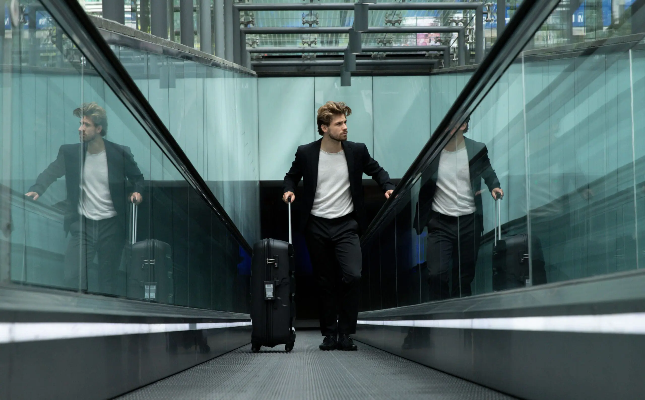 Man in pak met rolkoffer loopt door glazen roltrapgang in modern station of luchthaven.