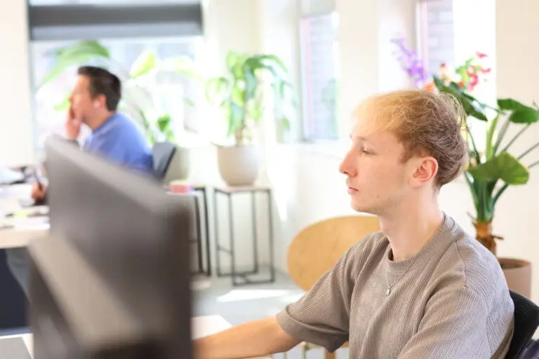 junior grafisch ontwerper van LOEKS werkt geconcentreerd achter een computerscherm in een lichte kantoorruimte met planten en een collega op de achtergrond.