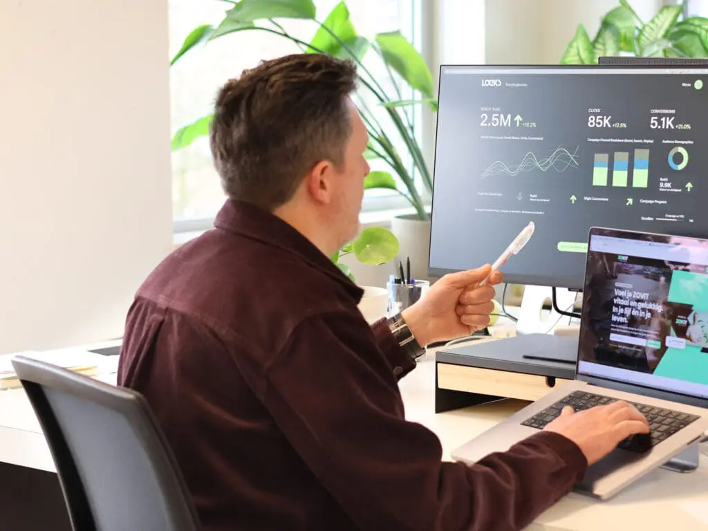 Eigenaar analyseert statistieken op een monitor en werkt op een laptop in een lichte kantooromgeving.