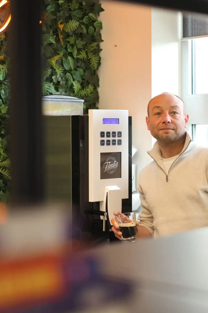 Eigenaar van Loeks staat bij een koffiemachine met een glas koffie, met een plantenwand op de achtergrond in een kantoorruimte.
