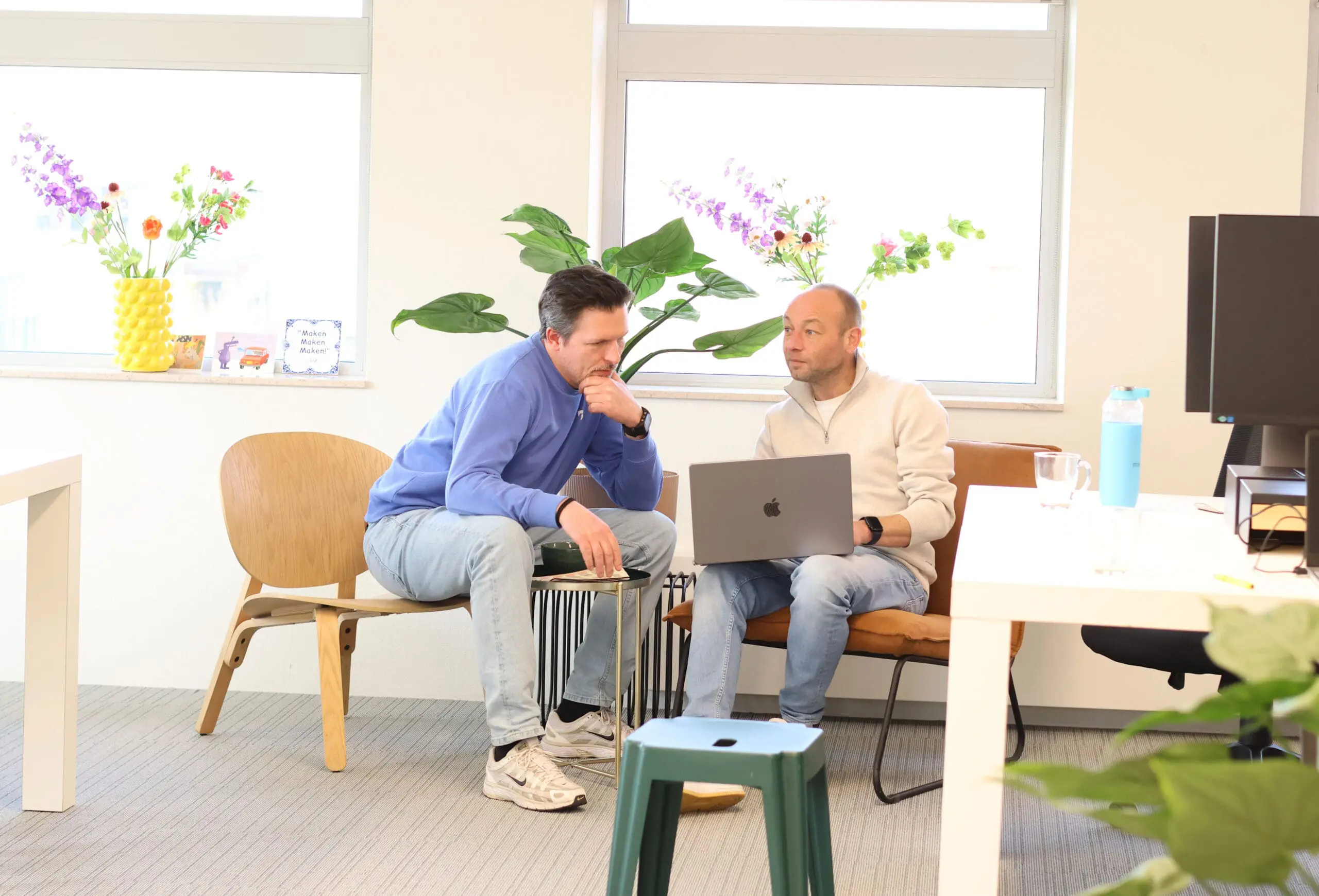 Twee collega’s van LOEKS bespreken iets op een laptop in een lichte kantoorruimte met planten op de achtergrond.