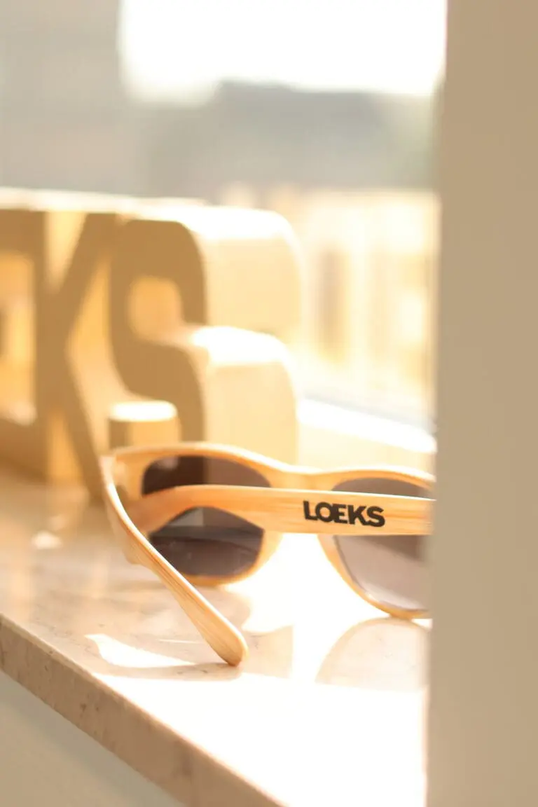 Houten zonnebril met “LOEKS” op pootje ligt op zonverlichte vensterbank, met wazige houten letters op achtergrond.