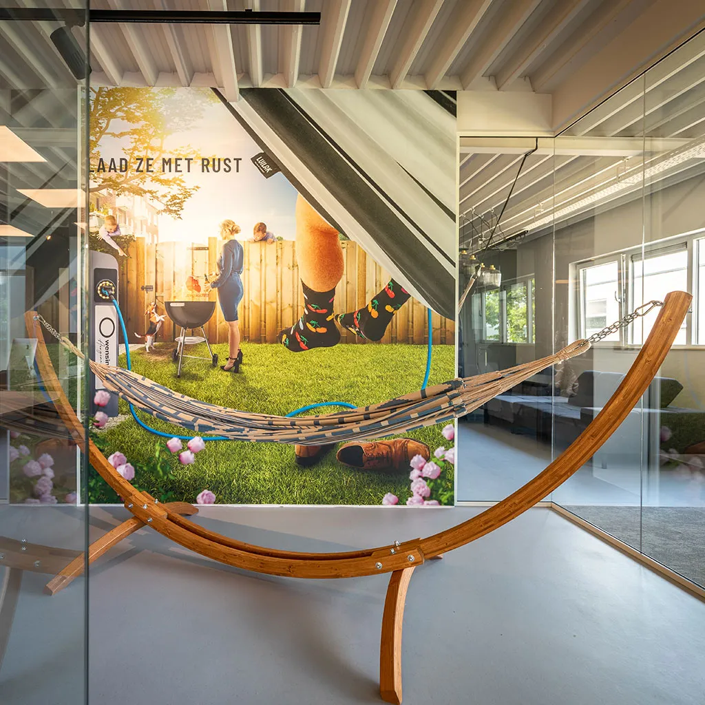 Hangmat op houten standaard in moderne ruimte met grote Luilak wandafbeelding van tuin met barbecue.