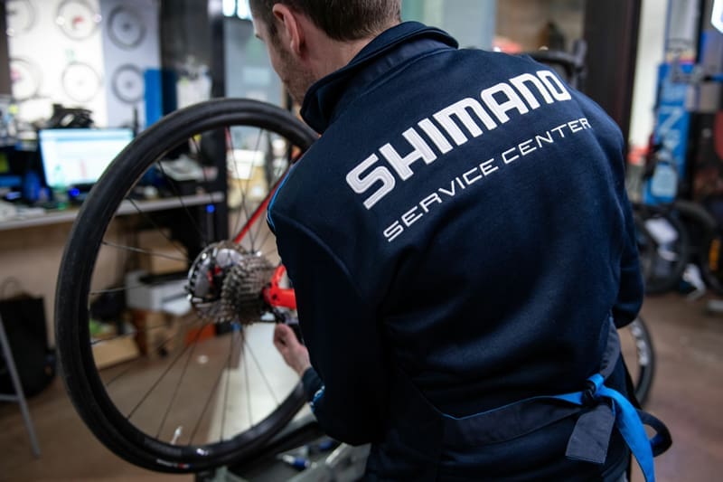 Fietsmonteur die bezig is met een fietsreparatie. Gekleed in een jack met de tekst Shimano Service Center