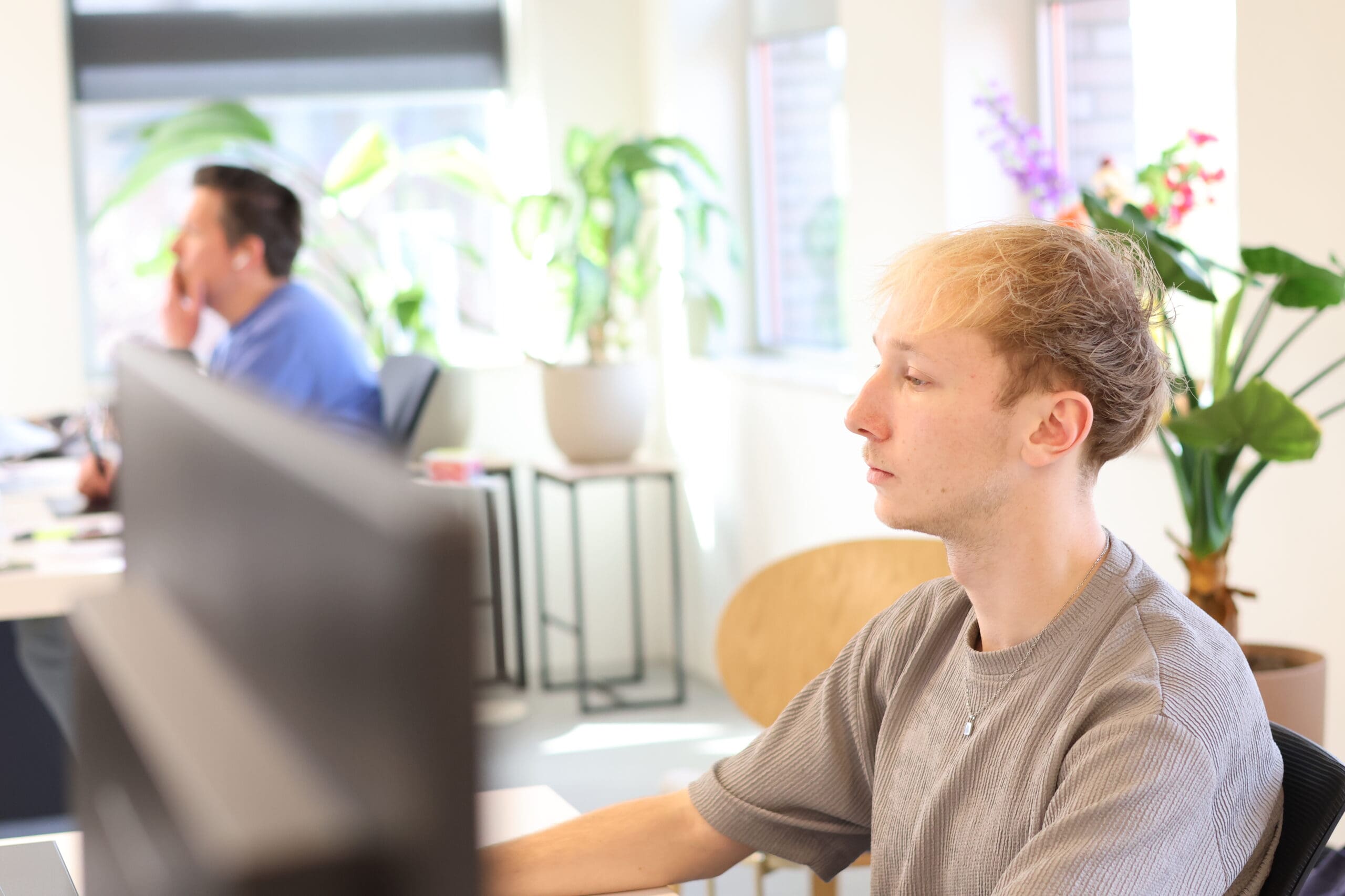 junior grafisch ontwerper van LOEKS werkt geconcentreerd achter een computerscherm in een lichte kantoorruimte met planten en een collega op de achtergrond.