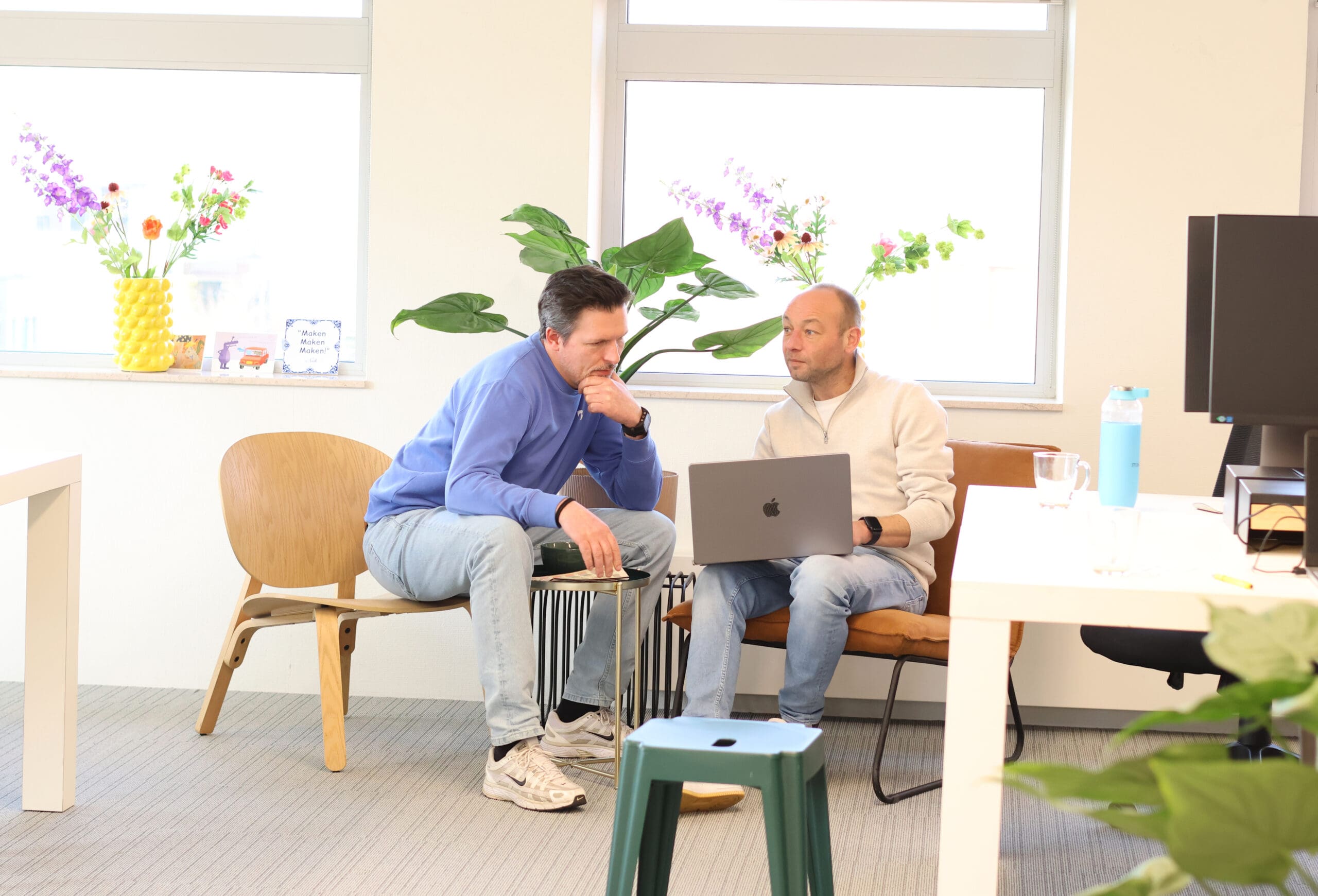 Twee collega’s van LOEKS bespreken iets op een laptop in een lichte kantoorruimte met planten op de achtergrond.